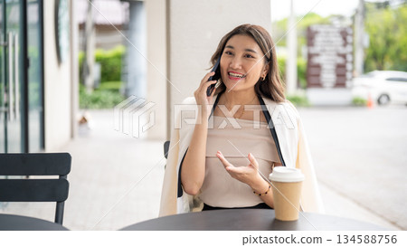 Asian woman smiling with explaining hand as talking on the phone with coffee cup on cafe black table Asian woman smiling with explaining hand as talking on the phone with coffee cup on cafe black table 134588756