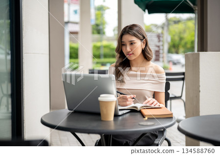 Long hair asian woman looking at laptop while holding pen over book with coffee aside at cafe table. Long hair asian woman looking at laptop while holding pen over book with coffee aside at cafe table. 134588760