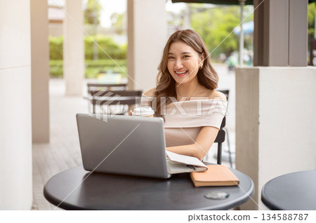 Long hair asian woman smiling happily while holding coffee and looking away from laptop on cafe table Long hair asian woman smiling happily while holding coffee and looking away from laptop on cafe table 134588787