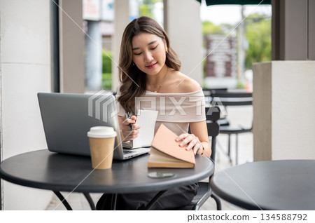 Long hair asian woman holding pen and opening a book with laptop and coffee cup aside at cafe table. 134588792