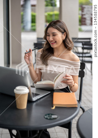 Long hair asian woman laughing as holding pen and book looking at laptop with coffee at cafe table. 134588795