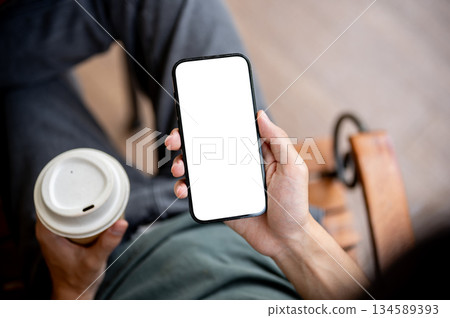 Close up of a hand holding blank screen smartphone reading news with a coffee cup on wooden bench. Close up of a hand holding blank screen smartphone reading news with a coffee cup on wooden bench. 134589393