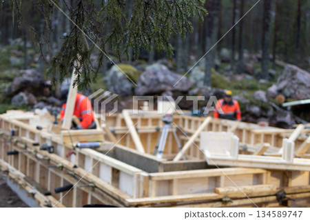 Construction workers in bright safety gear are assembling wooden framework for a building foundation in a forested area, showcasing teamwork and craftsmanship in a natural environment 134589747