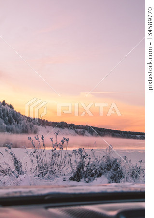 Winter landscape featuring snow-covered ground and frosty plants, with a serene lake reflecting the pastel colors of a sunset sky, creating a tranquil atmosphere in nature 134589770