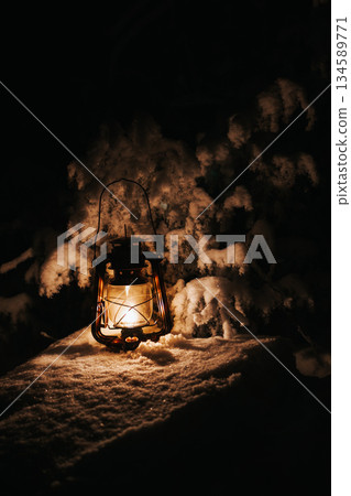 Vintage lantern glowing warmly on a snowy surface, surrounded by snow-covered branches, creating a cozy atmosphere in a winter night setting with enchanting light 134589771
