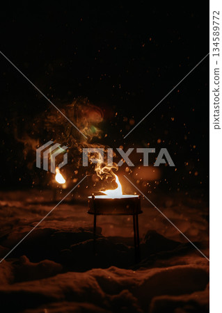 Flickering flame in a dark winter landscape, surrounded by falling snowflakes, creating a warm ambiance and inviting atmosphere for outdoor gatherings and cozy evenings 134589772