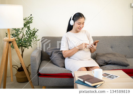 Pregnant asian woman wearing headphones listening to soothing music while sitting on sofa in house. 134589819