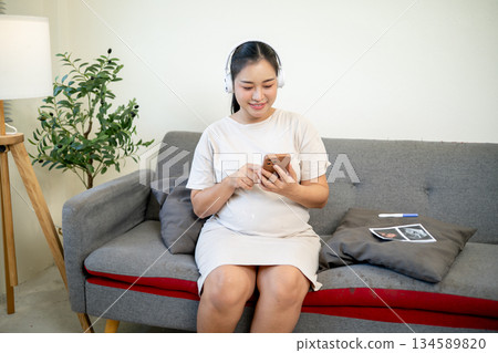 Pregnant asian woman wearing headphones listening to classical music while sitting on sofa in house. 134589820