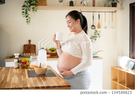 Pregnant asian woman holding a mug drinking water and touching her belly while standing in kitchen. Pregnant asian woman holding a mug drinking water and touching her belly while standing in kitchen. 134589823