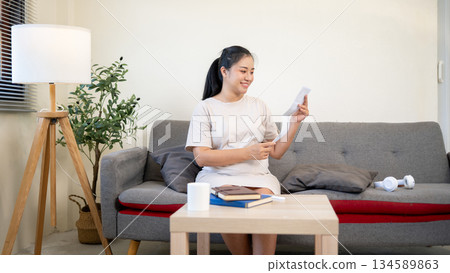 Pregnant asian woman is smiling as she looking at her ultrasound pictures while sitting on sofa in the house. 134589863