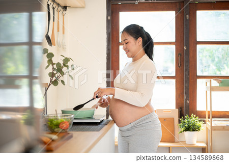 Side view of a pregnant asian woman standing and cooking a meal with a pan on stove in the kitchen. Side view of a pregnant asian woman standing and cooking a meal with a pan on stove in the kitchen. 134589868