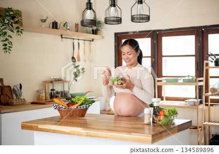 Pregnant asian woman smiling as holding glass bowl enjoy eating vegetables salad at kitchen counter. 134589896