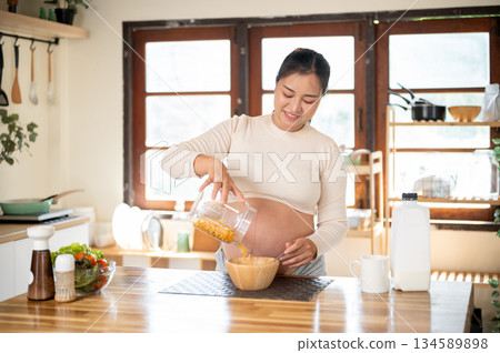 Pregnant asian woman pouring cereal in wooden bowl making herself a meal at kitchen cooking counter. 134589898