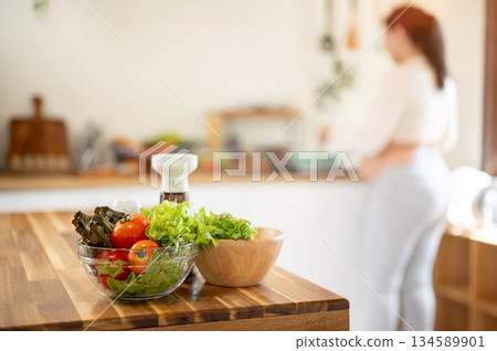 A glass bowl of vegetable on wooden cooking counter with blurred figure of pregnant woman in kitchen 134589901
