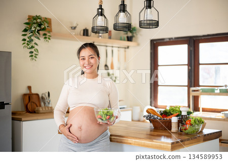 Pregnant asian woman holding vegetable salad glass bowl and touching her belly standing in kitchen. Pregnant asian woman holding vegetable salad glass bowl and touching her belly standing in kitchen. 134589953