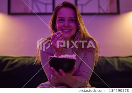 Female seated indoors eating popcorn and watching screen Female seated indoors eating popcorn and watching screen 134590378