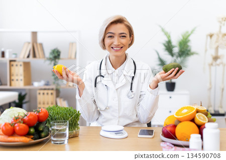 A smiling nutritionist in a white coat holds a lemon and avocado, surrounded by fresh produce, promoting healthy eating. A smiling nutritionist in a white coat holds a lemon and avocado, surrounded by fresh produce, promoting healthy eating. 134590708