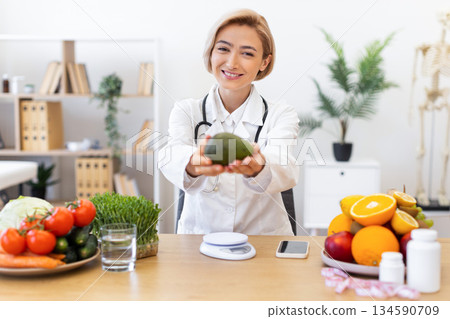 A smiling nutritionist in a white coat holds out an avocado, promoting healthy eating and wellness. A smiling nutritionist in a white coat holds out an avocado, promoting healthy eating and wellness. 134590709