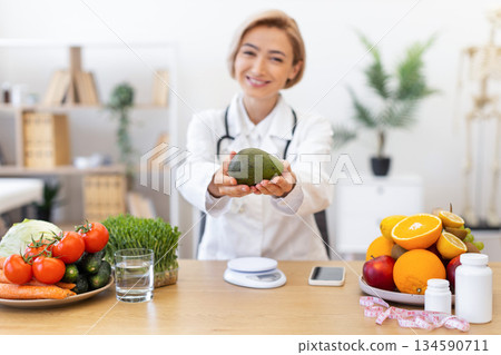 A smiling nutritionist holds an avocado, promoting healthy eating with fresh fruits and vegetables on a table. A smiling nutritionist holds an avocado, promoting healthy eating with fresh fruits and vegetables on a table. 134590711