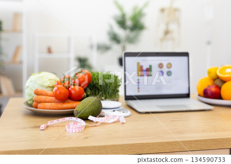 A laptop displays a diet plan next to fresh vegetables and fruits, with a measuring tape, suggesting a focus on healthy eating and weight management. 134590733