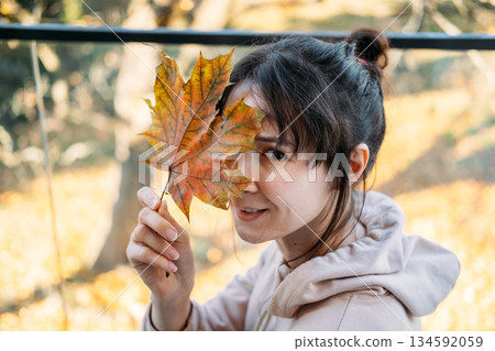 Young woman holds large maple leaf over eye in sunlit park. Seasonal transition, nature connection, mindful living, outdoor wellness. 134592059