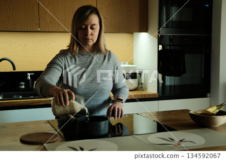 Woman stands in modern kitchen pouring rice from glass jar into small pot on induction stove. Vegan cooking, plant-based diet, healthy ingredients, natural seasoning. 134592067