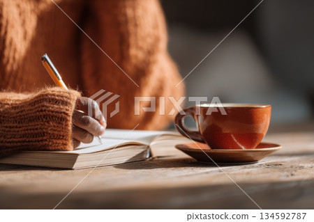 A person writes in a notebook while holding a pen. A cup of coffee sits nearby on a saucer. The setting is relaxed and warm 134592787