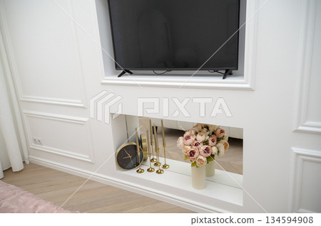 Bedroom interior, white decorative wall with TV, clock, vase of flowers, candles. 134594908