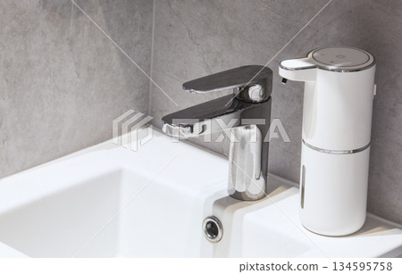 Close-up of a chrome faucet and white touchless liquid soap dispenser in a minimalist bathroom interior. The concept of hygiene, cleanliness, modern smart home technology, and hand washing. Close-up of a chrome faucet and white touchless liquid soap dispenser in a minimalist bathroom interior. The concept of hygiene, cleanliness, modern smart home technology, and hand washing. 134595758