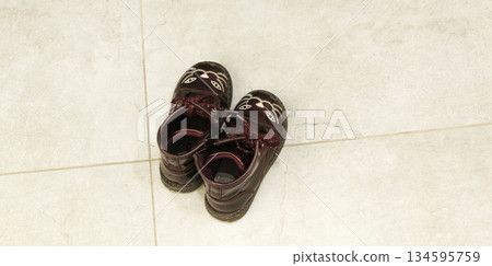 A pair of burgundy leather children's boots with a cute cat design, standing on light-colored tiles. Top view, concept of childhood, coming home, fashion for kids, and family comfort in the interior. A pair of burgundy leather children's boots with a cute cat design, standing on light-colored tiles. Top view, concept of childhood, coming home, fashion for kids, and family comfort in the interior. 134595759