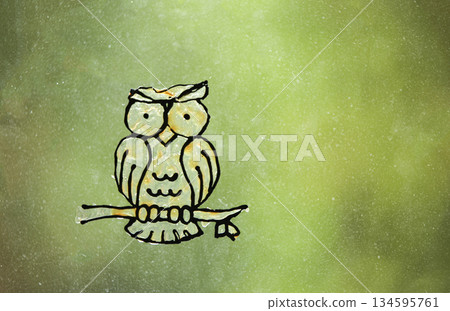 A homemade sticker or drawing of a yellow owl attached to a transparent pane of glass. A blurred green background of a garden, the concept of children's creativity, DIY crafts, and summer vacation. 134595761