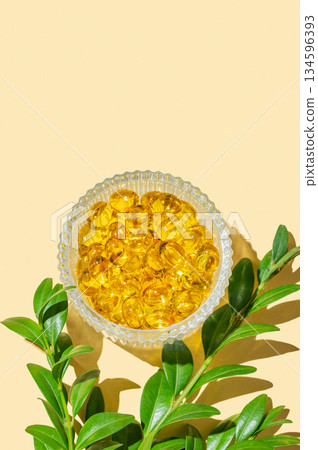 Yellow softgel vitamins capsules in a bowl on a bright background with green leaves. Yellow softgel vitamins capsules in a bowl on a bright background with green leaves. 134596393