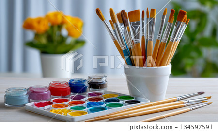 A vibrant array of art supplies is on a table, A palette with various watercolors and a set of paintbrushes in a holder 134597591