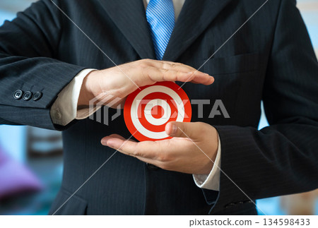 A man in a business suit holds a target in his palms. A candidate for a position. Setting a goal to achieve. Career aspiration, recruitment decision. Evaluation process and personal responsibility. 134598433