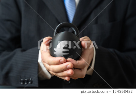 A heavy burden of debt in the hands of a european businessman. A weight with the euro symbol in the hands of a businessman. Rising interest costs create risks. Weight of obligations slows development. 134598482