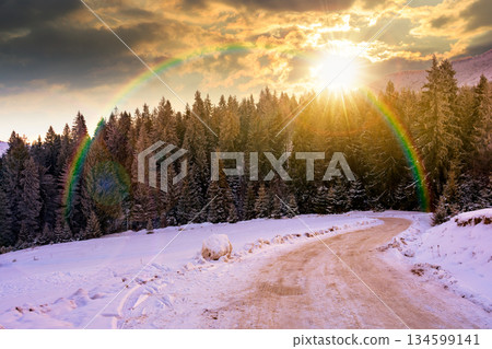 winter landscape with snow covered country road winding through fir forest in mountains at sunset. alpine countryside of in evening light. timeless nostalgia book cover with rainbow 134599141