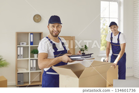 Two professional friendly movers from cardboard boxes unpacking office items in new location. 134601736