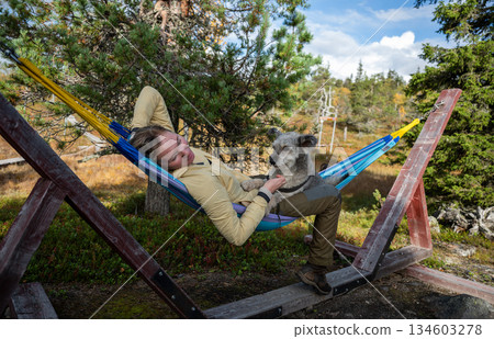Woman relaxing in hammock with dog wearing outdoor clothing in forest nature Woman relaxing in hammock with dog wearing outdoor clothing in forest nature 134603278