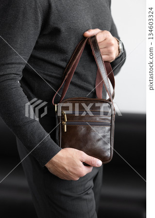Stylish man in charcoal knitwear and trousers holds a dark brown vertical leather messenger bag against a white studio backdrop Stylish man in charcoal knitwear and trousers holds a dark brown vertical leather messenger bag against a white studio backdrop 134603324