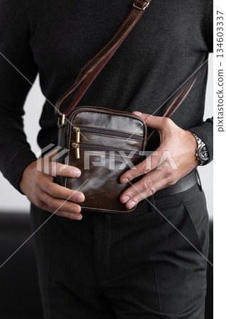 Stylish man in charcoal knitwear and trousers holds a dark brown vertical leather messenger bag against a white studio backdrop Stylish man in charcoal knitwear and trousers holds a dark brown vertical leather messenger bag against a white studio backdrop 134603337