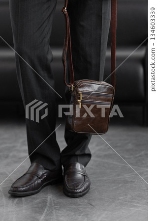 Stylish man in charcoal knitwear and trousers holds a dark brown vertical leather messenger bag against a white studio backdrop Stylish man in charcoal knitwear and trousers holds a dark brown vertical leather messenger bag against a white studio backdrop 134603339