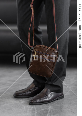 Stylish man in charcoal knitwear and trousers holds a dark brown vertical leather messenger bag against a white studio backdrop Stylish man in charcoal knitwear and trousers holds a dark brown vertical leather messenger bag against a white studio backdrop 134603343