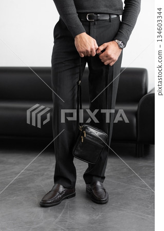 Male model in a charcoal knit crewneck and tailored trousers wearing a black leather vertical messenger bag against a white studio background. Male model in a charcoal knit crewneck and tailored trousers wearing a black leather vertical messenger bag against a white studio background. 134603344