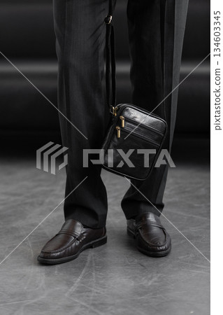 Male model in a charcoal knit crewneck and tailored trousers wearing a black leather vertical messenger bag against a white studio background. Male model in a charcoal knit crewneck and tailored trousers wearing a black leather vertical messenger bag against a white studio background. 134603345