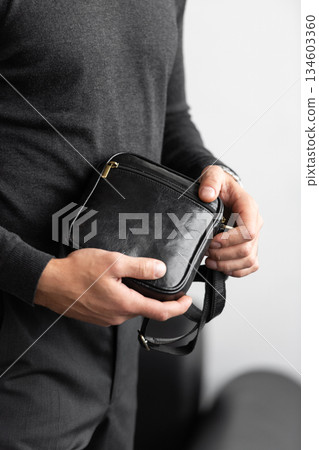 Male model in a charcoal knit crewneck and tailored trousers wearing a black leather vertical messenger bag against a white studio background. Male model in a charcoal knit crewneck and tailored trousers wearing a black leather vertical messenger bag against a white studio background. 134603360