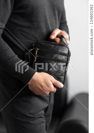 Male model in a charcoal knit crewneck and tailored trousers wearing a black leather vertical messenger bag against a white studio background. Male model in a charcoal knit crewneck and tailored trousers wearing a black leather vertical messenger bag against a white studio background. 134603362