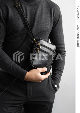 Male model in a charcoal knit crewneck and tailored trousers wearing a black leather vertical messenger bag against a white studio background. Male model in a charcoal knit crewneck and tailored trousers wearing a black leather vertical messenger bag against a white studio background. 134603370