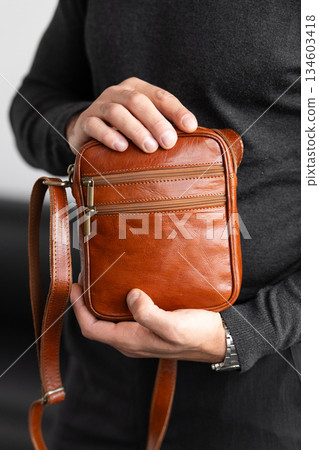 Male model in a charcoal knit crewneck and tailored trousers wearing a cognac leather vertical messenger bag against a white studio background. 134603418
