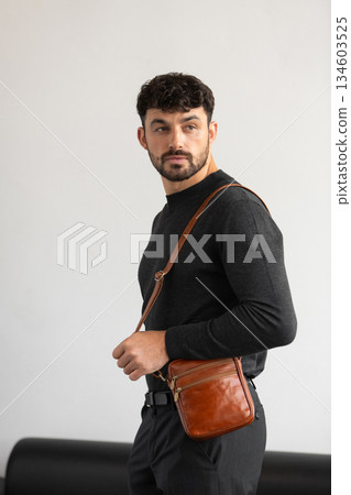 A stylish man in a dark grey crew neck sweater poses with a cognac leather crossbody bag against a minimalist white studio background. 134603525
