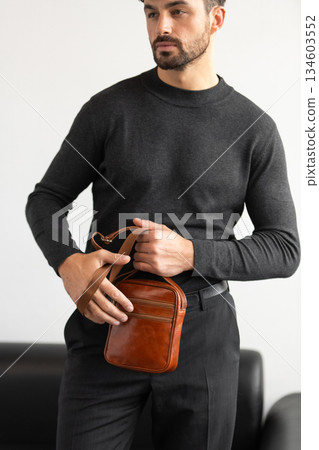 A stylish man in a dark grey crew neck sweater poses with a cognac leather crossbody bag against a minimalist white studio background. A stylish man in a dark grey crew neck sweater poses with a cognac leather crossbody bag against a minimalist white studio background. 134603552